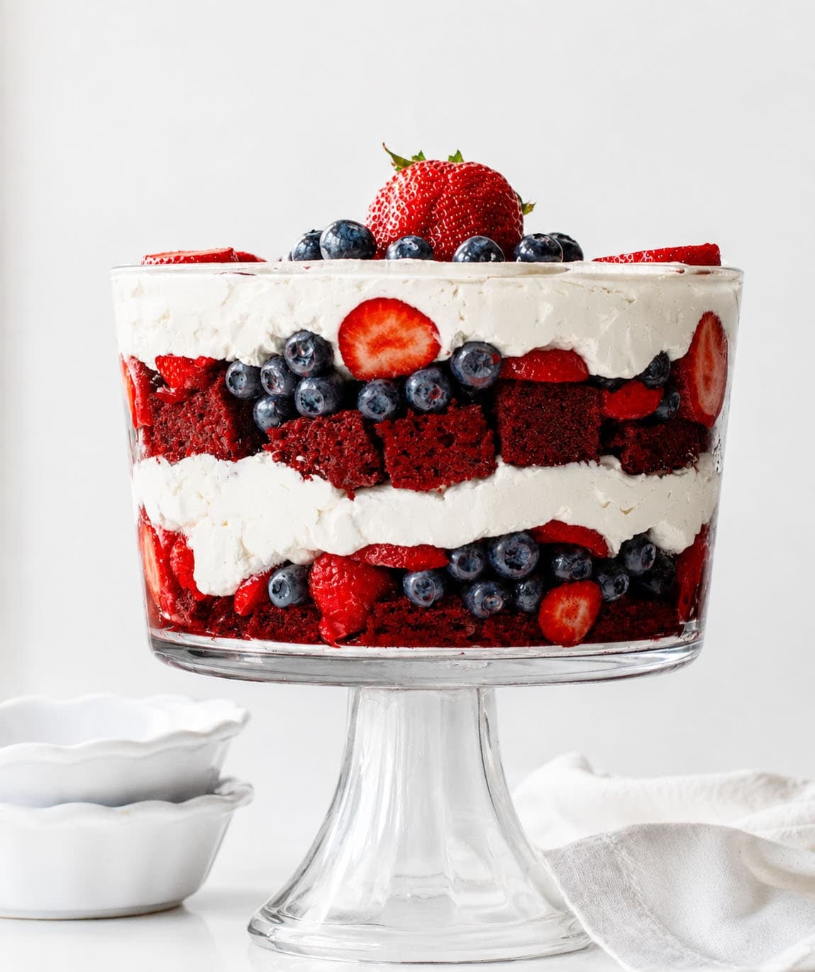 Layered Red Velvet Berry Trifle with Fresh Berries and Whipped Cream Looking at a Red Velvet Berry Trifle on a white counter top with white dishes and towel.