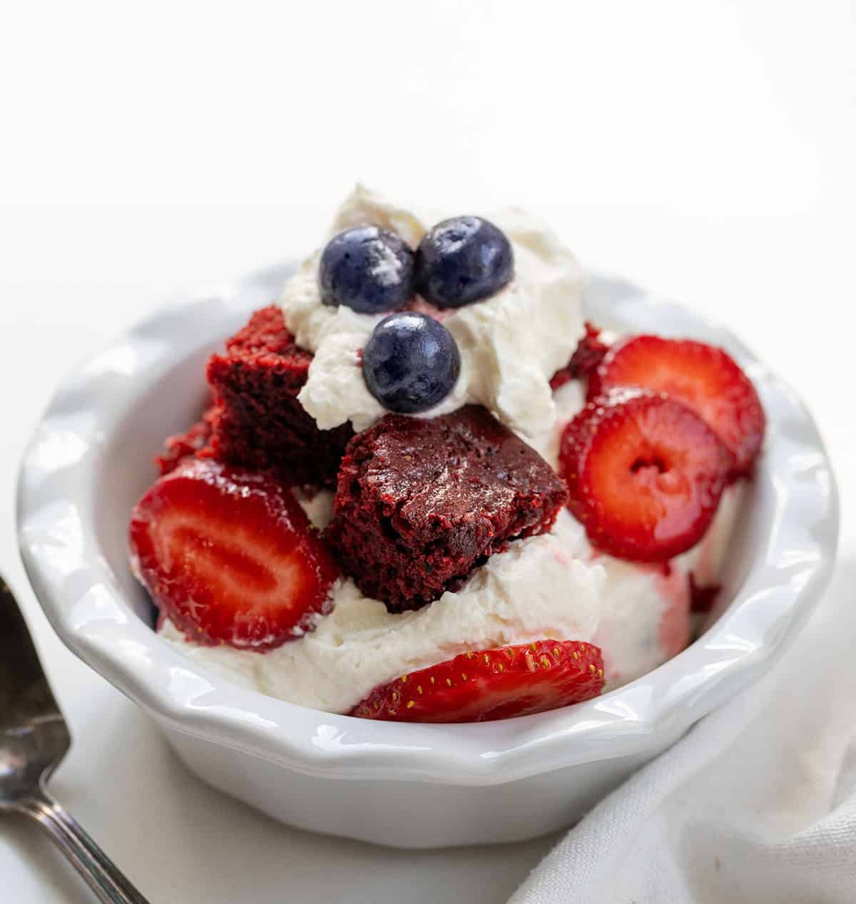 Layered Red Velvet Berry Trifle with Fresh Berries and Whipped Cream Bowl of Red Velvet Berry Trifle on a white table with white towel and spoon.