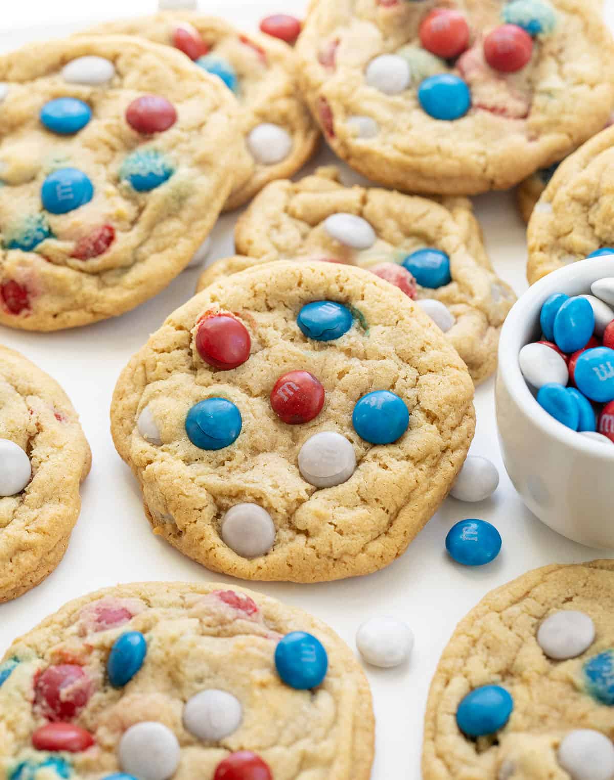 Close up of Red, White, and Blue M&M Cookies on a white table with a white bowl of M&M's next to the cookies.