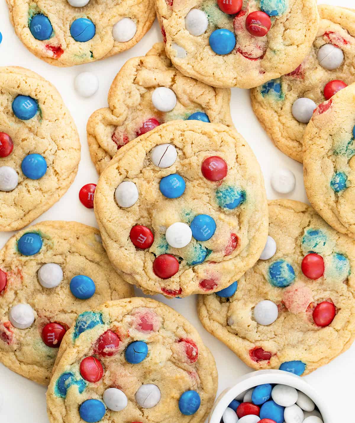 Close up of Red, White, and Blue M&M Cookies on a white table with a white bowl of M&M's next to the cookies.