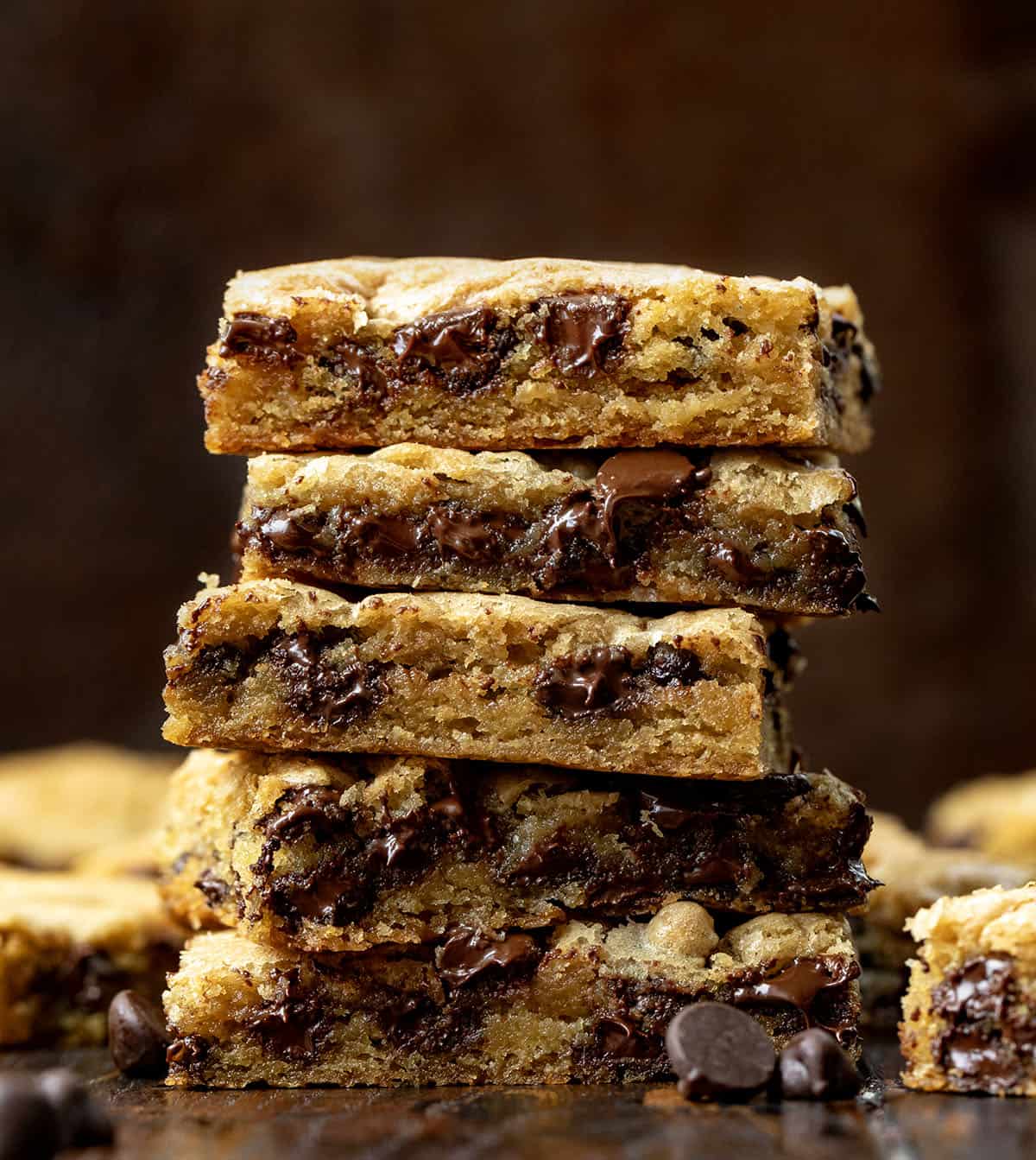These Sheet Pan Chocolate Chip Cookies are an easy way to make a big batch of soft, chewy chocolate chip cookies in one pan. Thick, buttery, and loaded with chocolate chips, they bake up golden on the edges with a soft center in every bite. Stack of cut Sheet Pan Chocolate Chip Cookies on a wooden table surrounded by more sheet pan cookie bars.