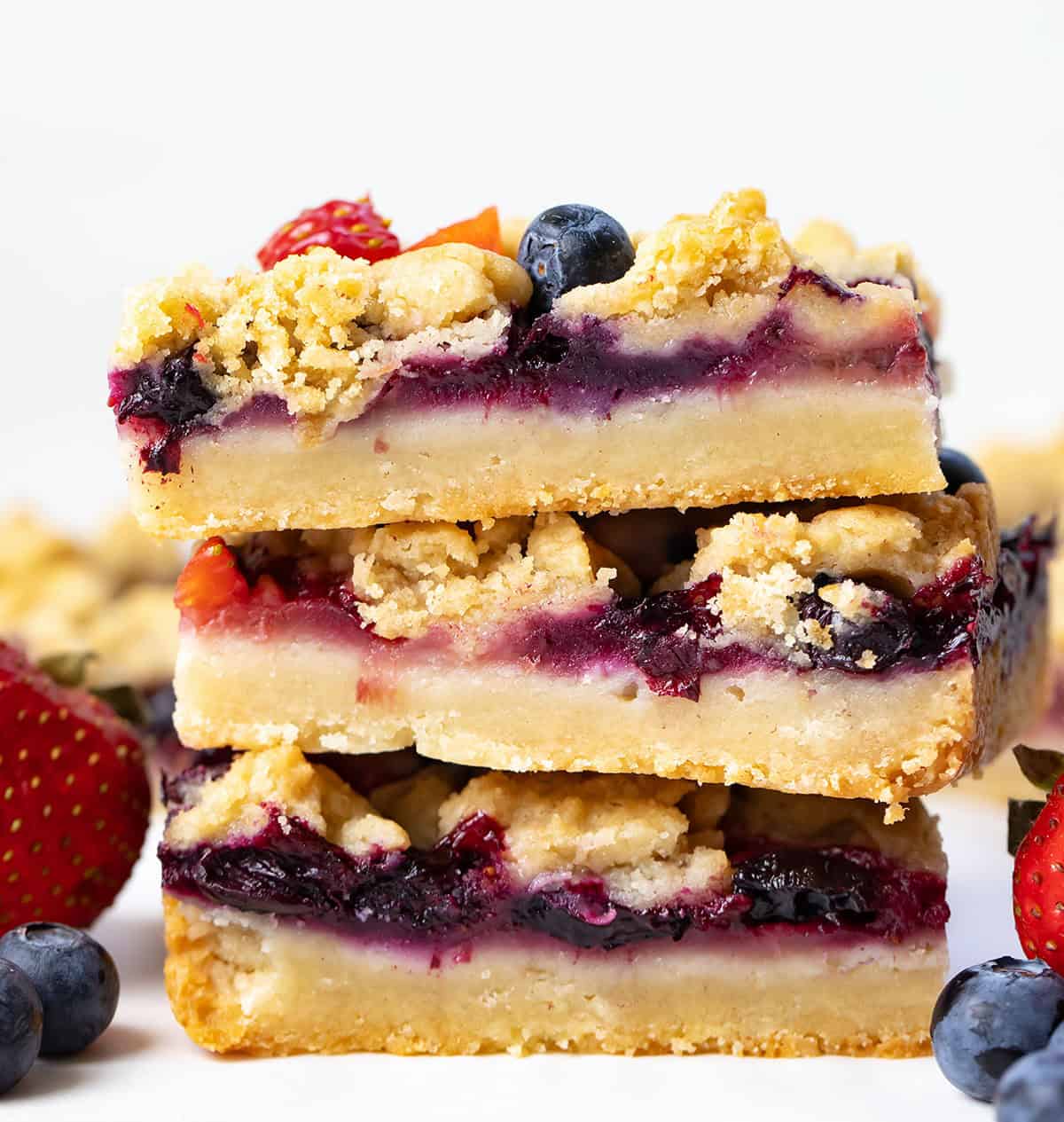 Stack of Strawberry Blueberry Crumble Bars on a white table with fresh blueberries and strawberries.