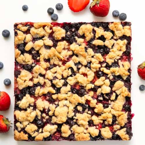 Strawberry Blueberry Crumble Bars fresh from the oven on a white table with fresh blueberries and strawberries.