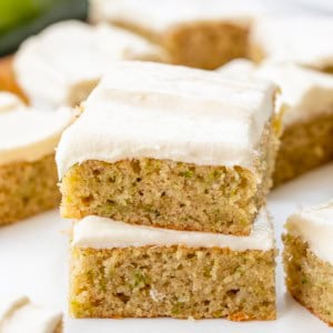 Zucchini Bars covered in cream cheese frosting stacked on a white table and surrounded by more bars.