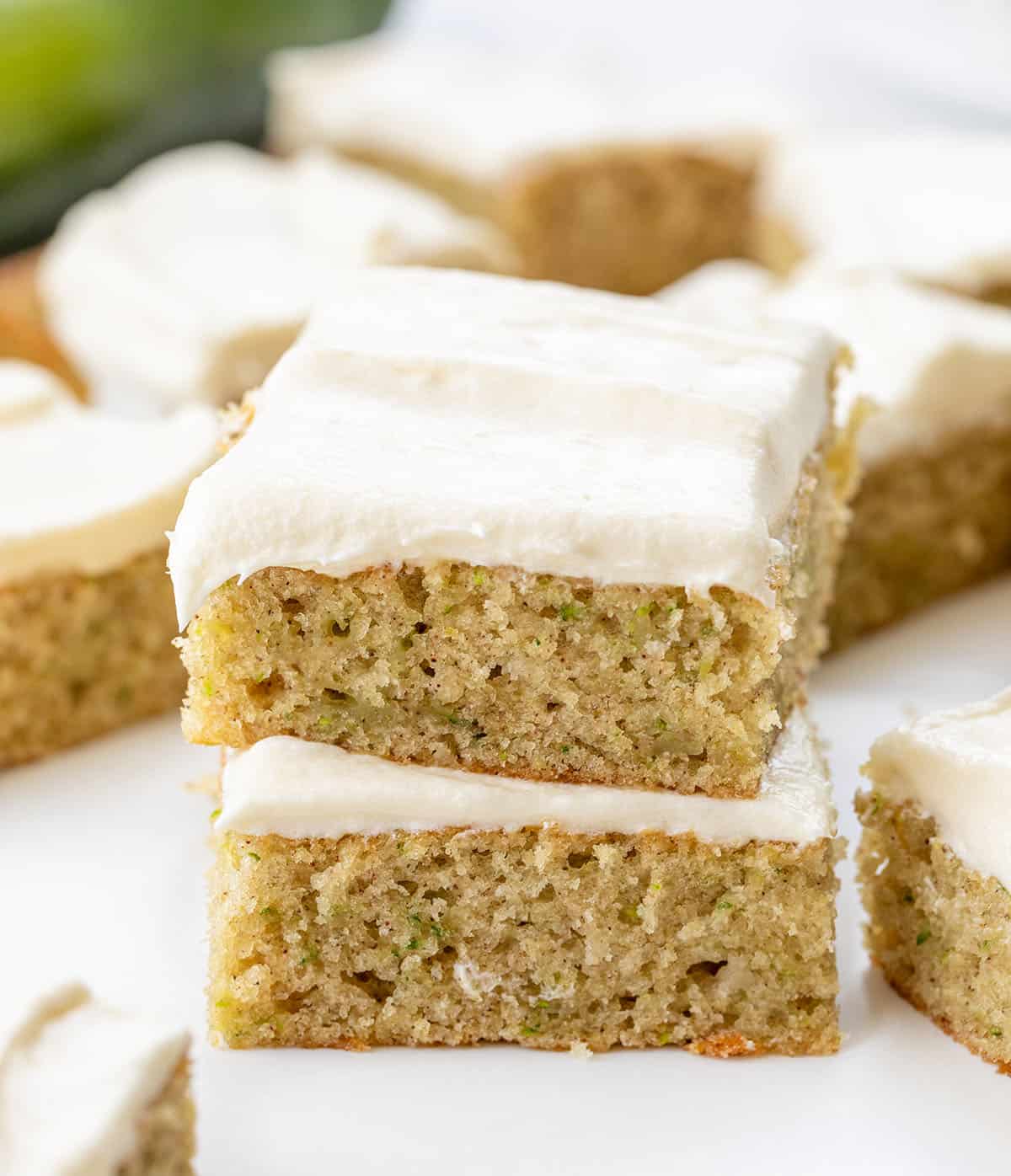Zucchini Bars covered in cream cheese frosting stacked on a white table and surrounded by more bars.