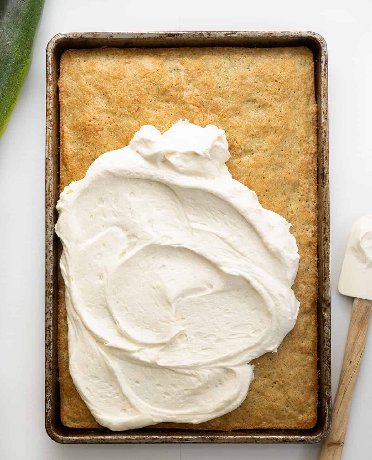 Covering a pan of Zucchini Bars in cream cheese frosting with the pan on a white table. 