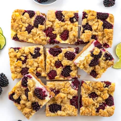 Blackberry Lime Cheesecake Bars cut into squares surrounded by limes and blackberries on a white table.