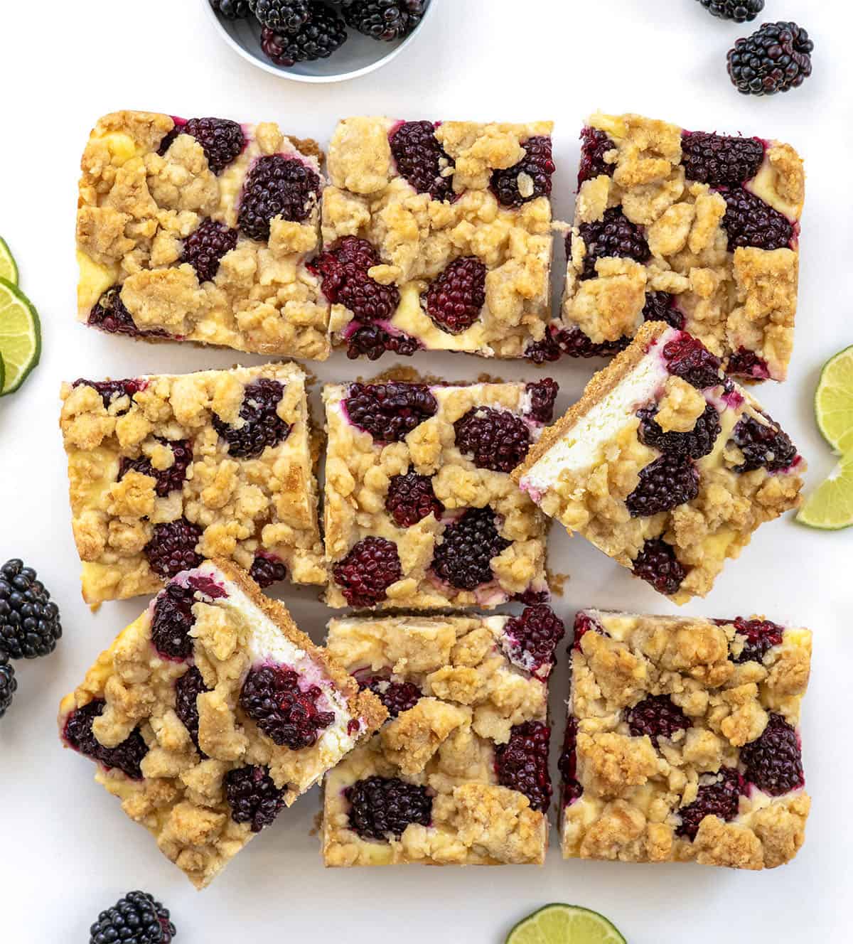 Blackberry Lime Cheesecake Bars cut into squares surrounded by limes and blackberries Blackberry Lime Cheesecake Bars cut into squares surrounded by limes and blackberries on a white table.
