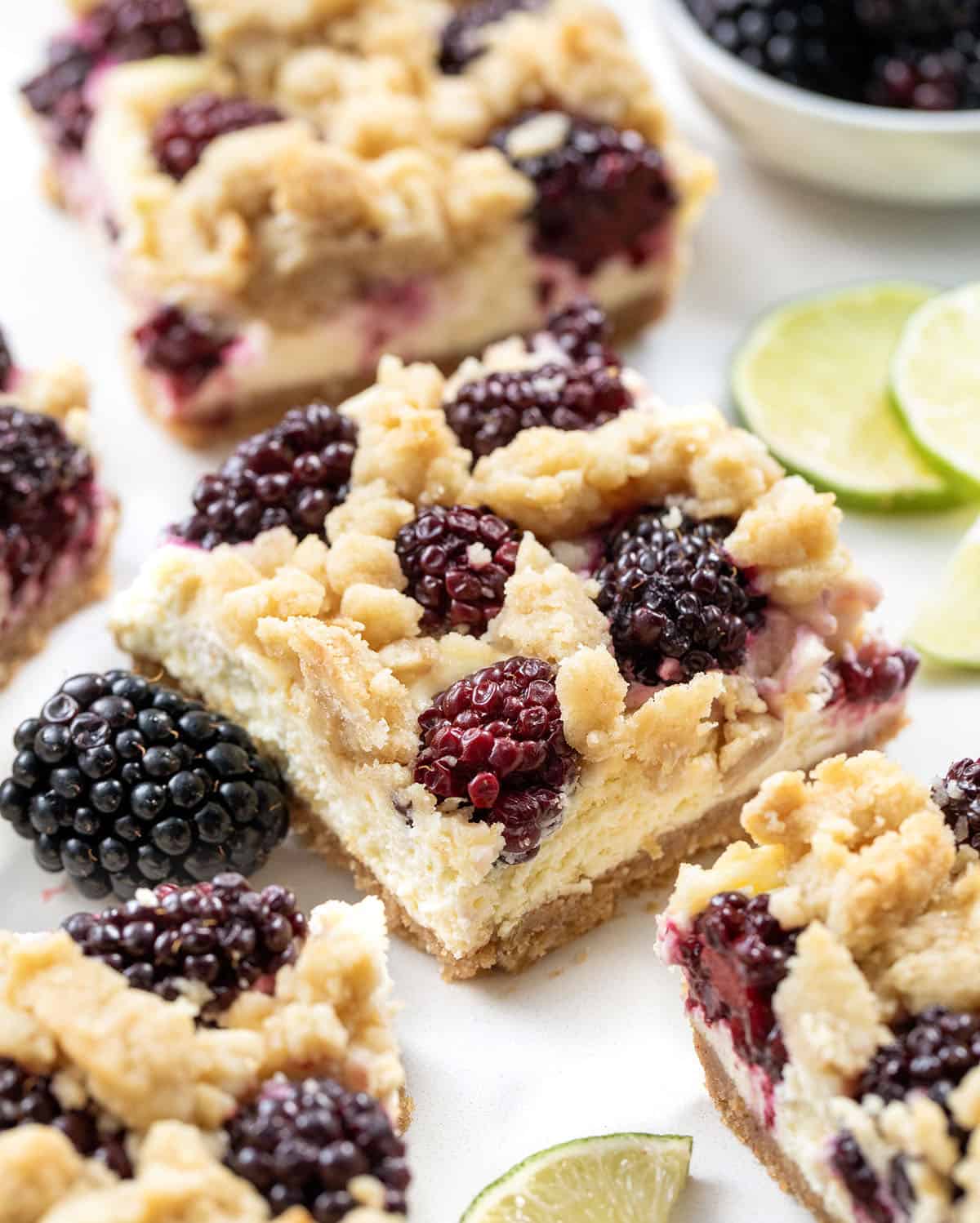 Close up of Blackberry Lime Cheesecake Bar surrounded by limes and blackberries.