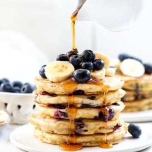 Pouring syrup over a stack of Blueberry Banana Buttermilk Pancakes.