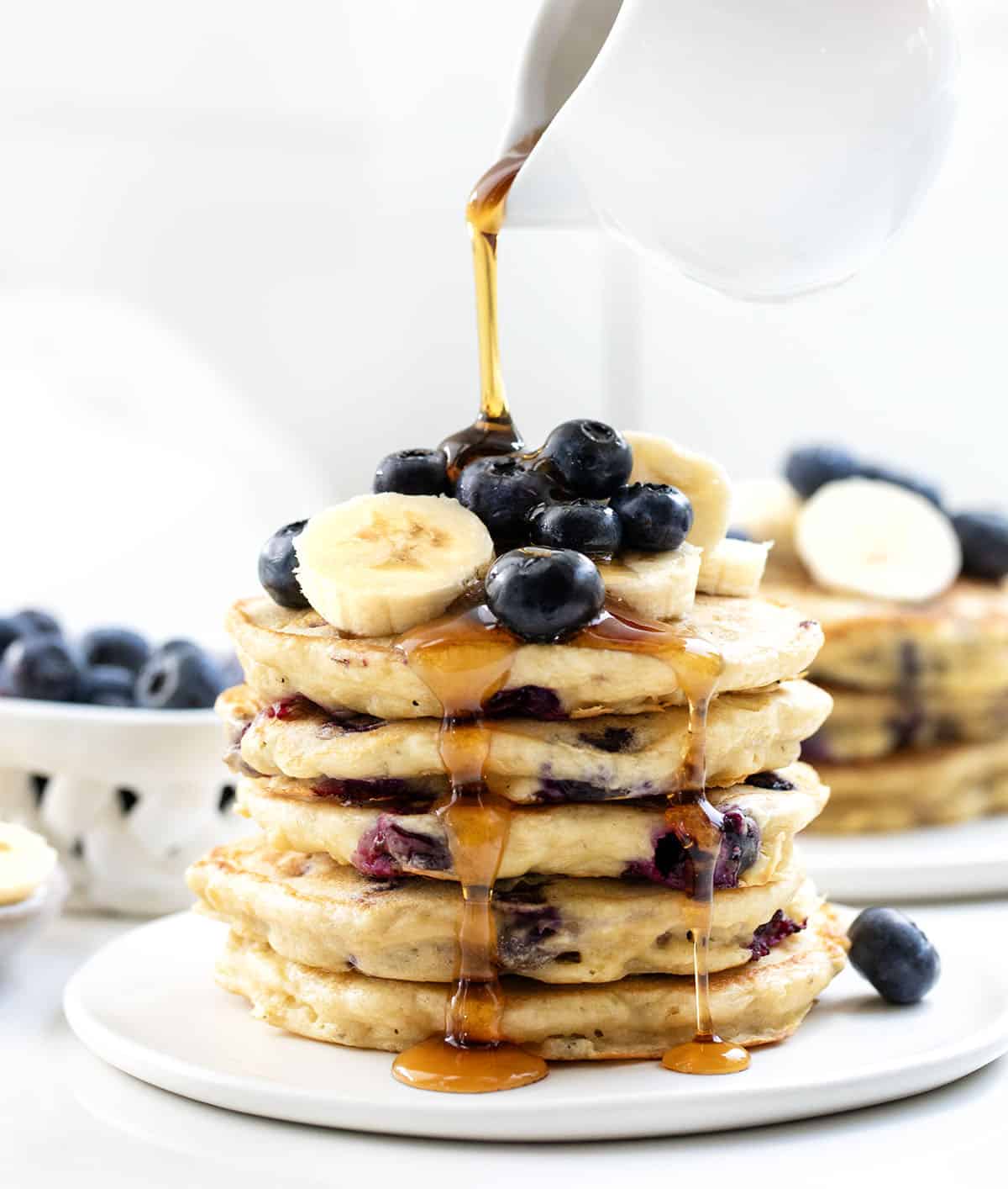 Pouring syrup over a stack of Blueberry Banana Buttermilk Pancakes.