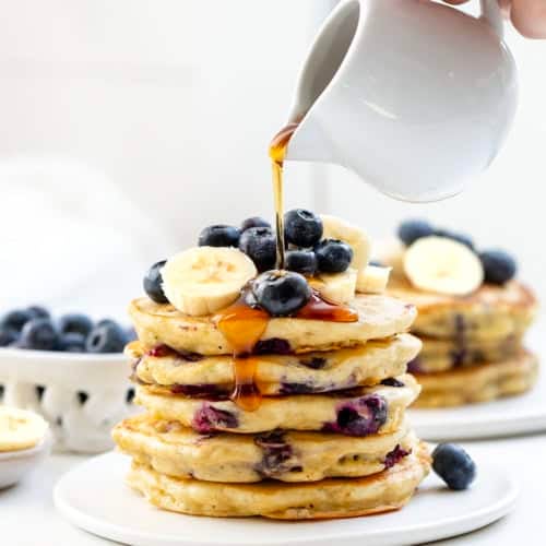 Pouring syrup over a stack of Blueberry Banana Buttermilk Pancakes.