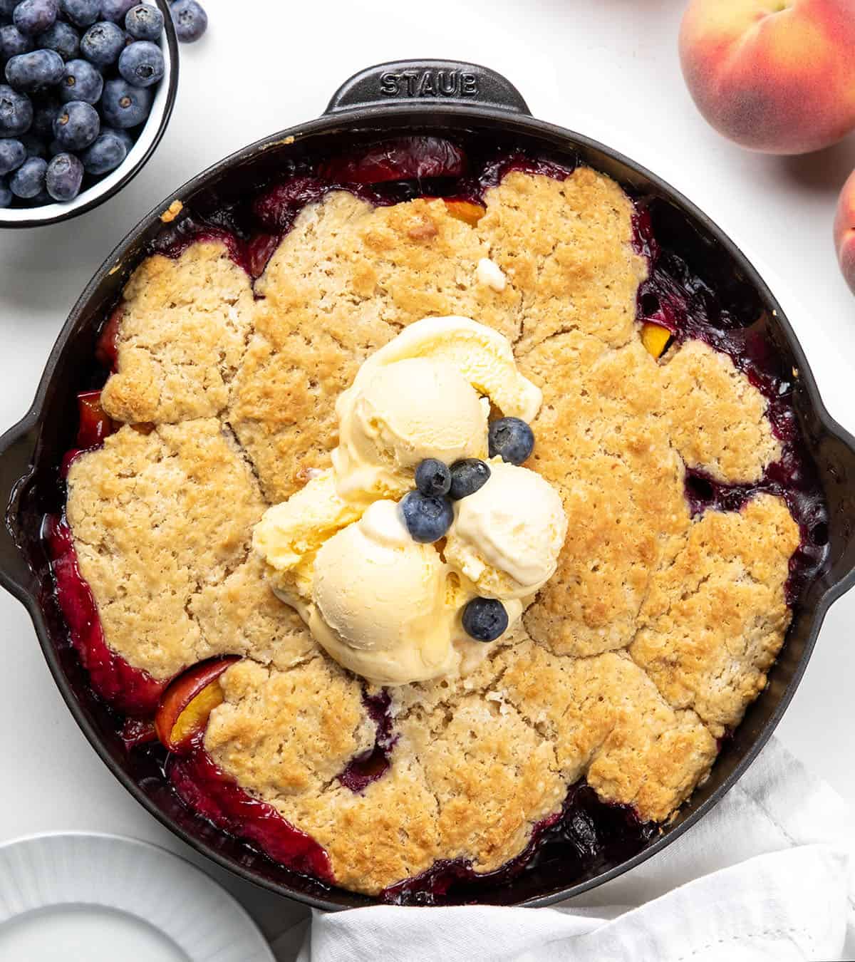 Skillet of Blueberry Peach Cobbler on a white table with blueberries and peaches.