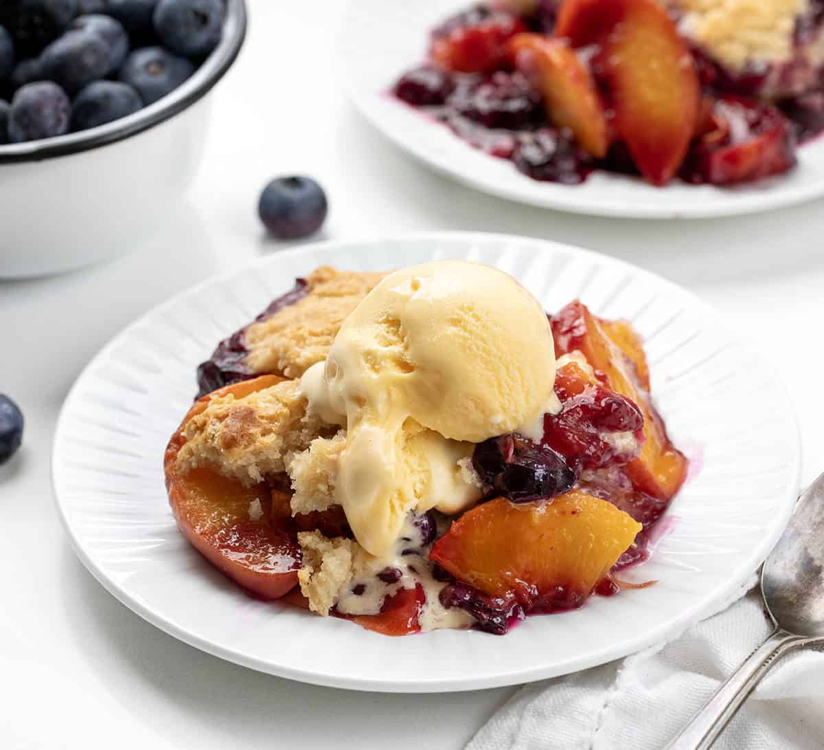 White plates of Blueberry Peach Cobbler on a white table with fresh blueberries and peaches.