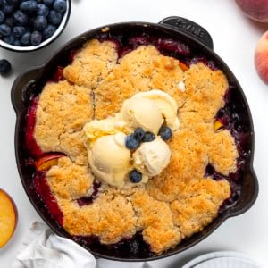 Skillet of Blueberry Peach Cobbler on a white table with blueberries and peaches.