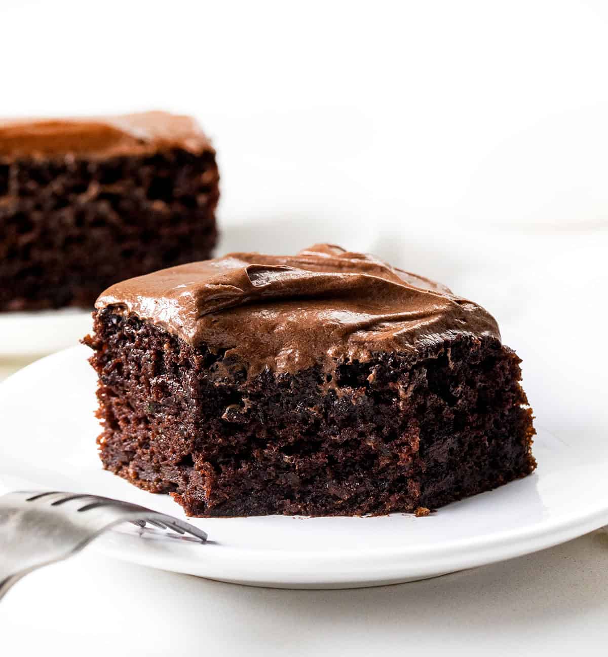 Slice of Chocolate Zucchini Snack Cake on a white plate with a bite removed and fork resting on a plate Slice of Chocolate Zucchini Snack Cake on a white plate with a bite removed and fork resting on a plate.