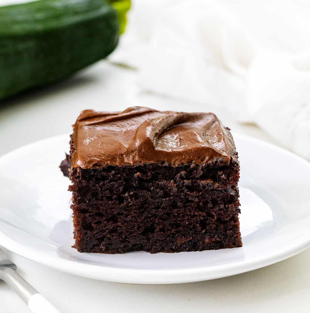 Chocolate Zucchini Snack Cake on a white plate on a white table with zucchini in the background Chocolate Zucchini Snack Cake on a white plate on a white table with zucchini in the background.