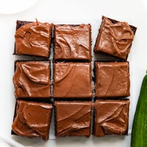 Chocolate Zucchini Snack Cake cut into squares on a white table with a zucchini.
