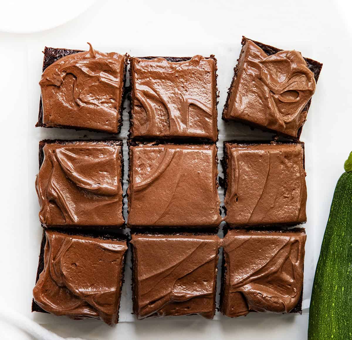 Chocolate Zucchini Snack Cake cut into squares on a white table with a zucchini.