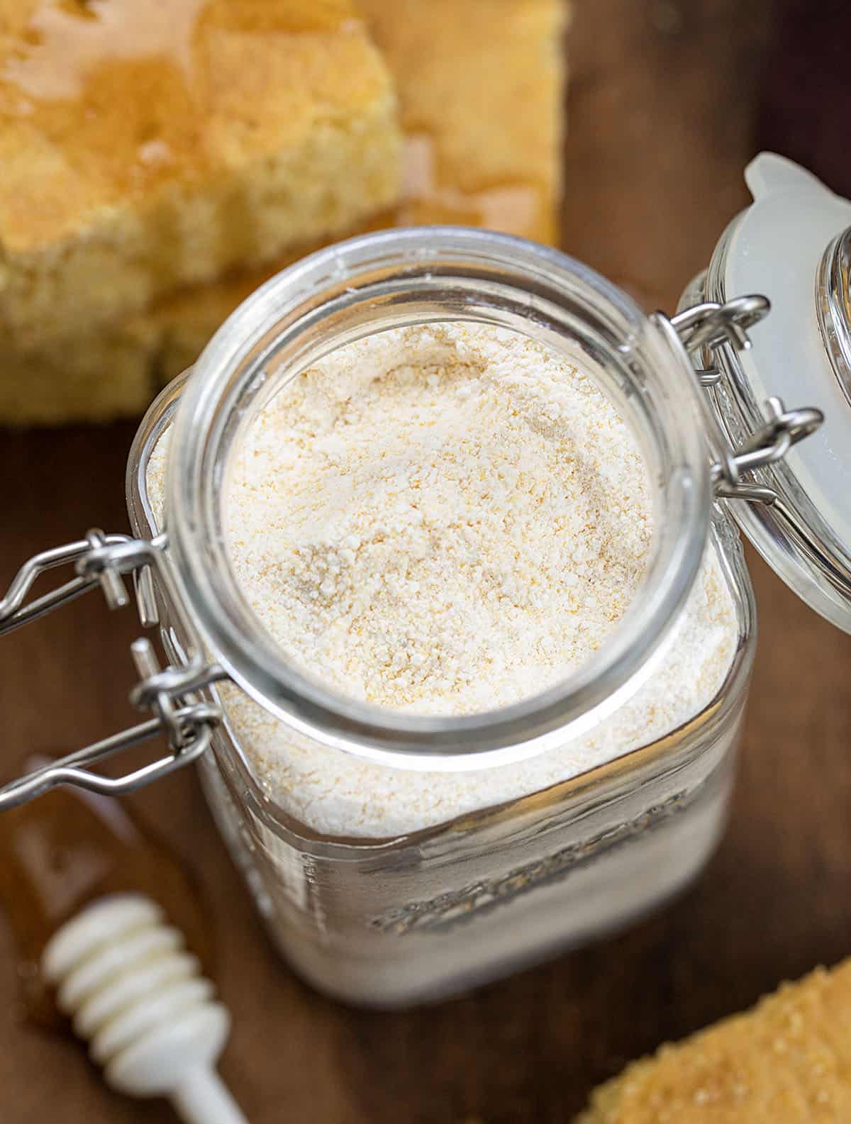 Close up on the open jar of Homemade Cornbread Mix on a wooden table with cornbread and honey.