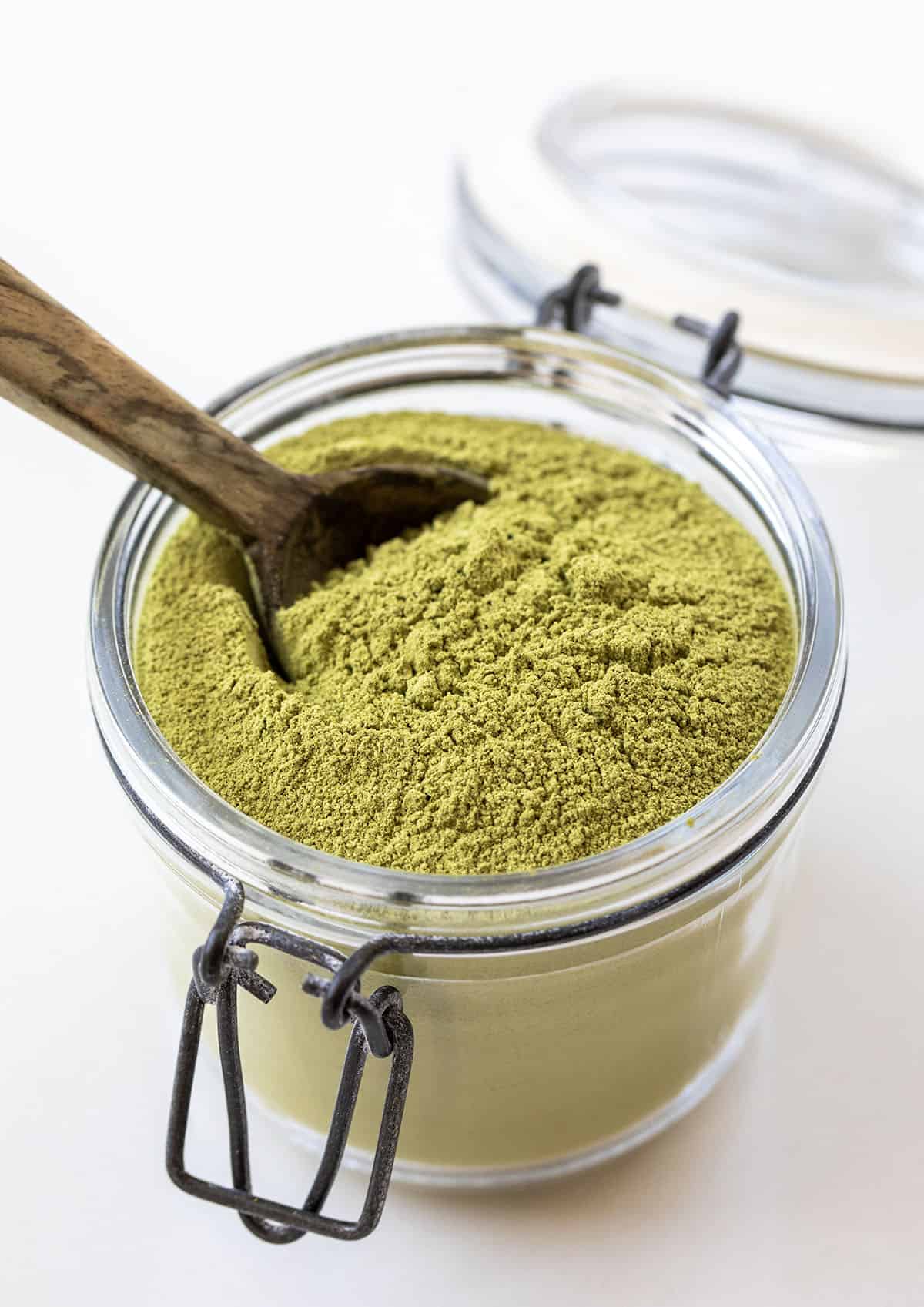 Jar of Matcha Latte Mix on a white table with a wooden spoon in it Jar of Matcha Latte Mix on a white table with a wooden spoon in it.