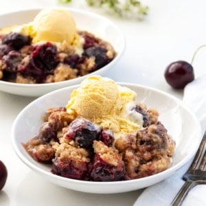 Bowls of Snickerdoodle Cherry Cobbler on a white table with ice cream and cherries.