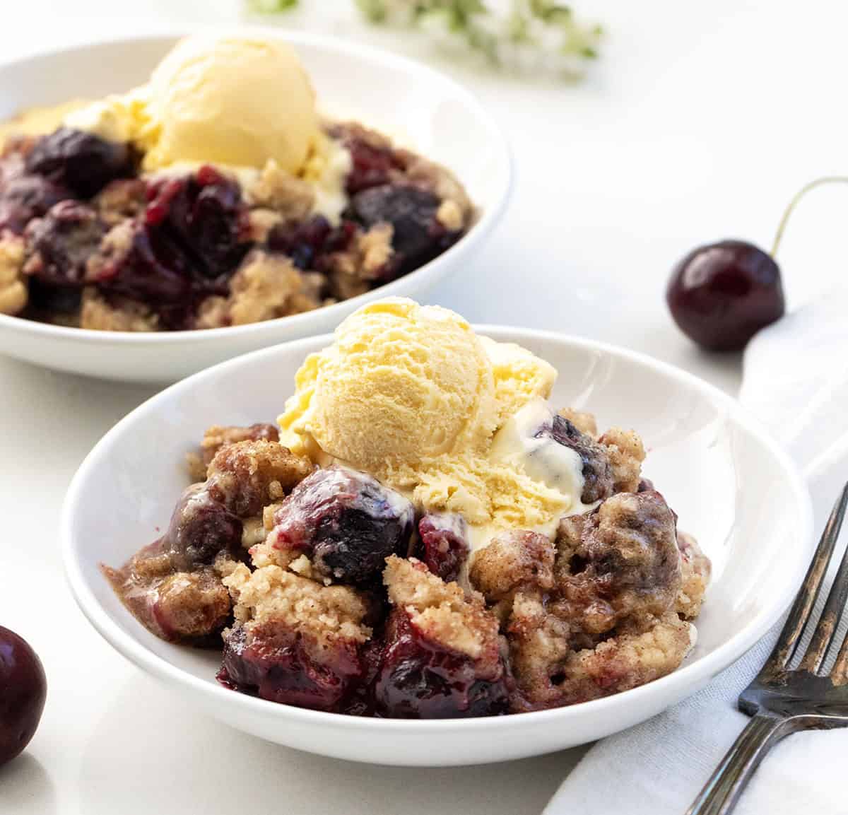 Bowls of Snickerdoodle Cherry Cobbler on a white table with ice cream and cherries.