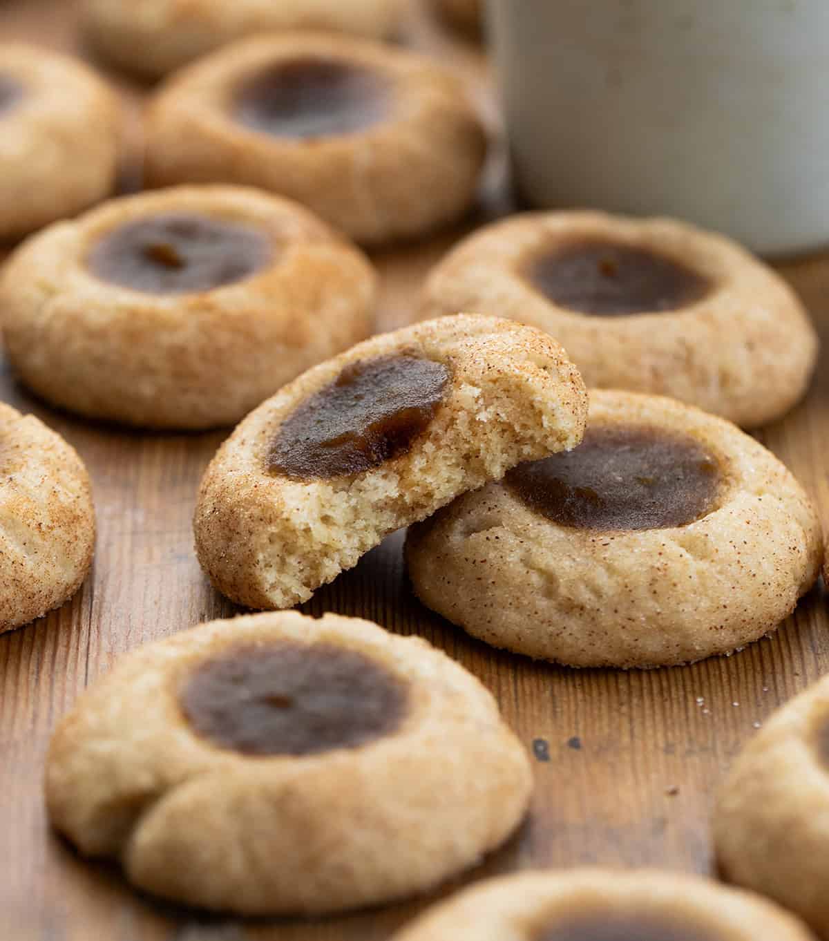 One Apple Butter Thumbprint Cookie has a bite taken out of it and is surrounded by more apple butter thumbprint cookies on a wooden table.