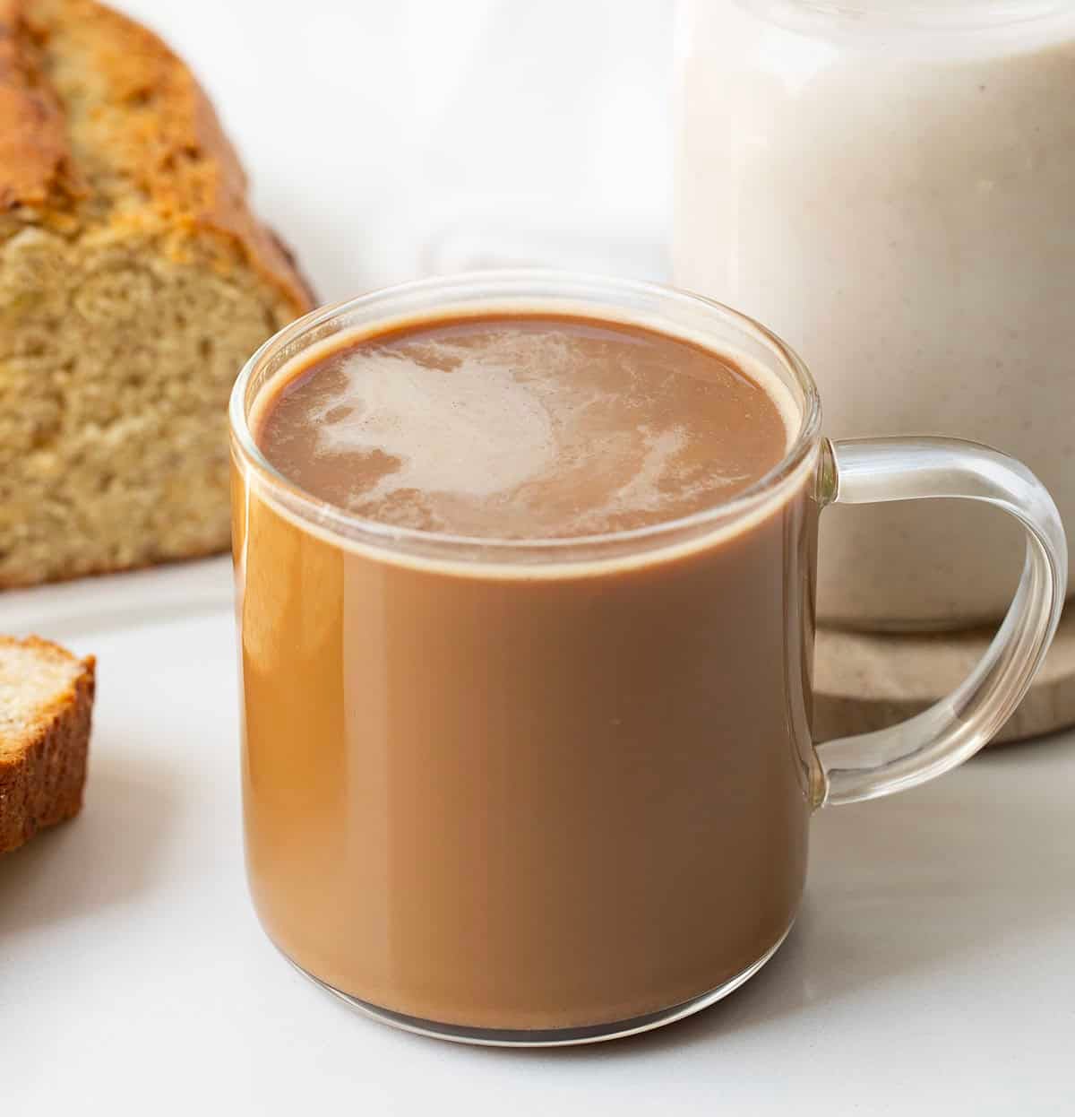 Glass cup of coffee with Banana Bread Coffee Creamer on it and banana bread in the background.