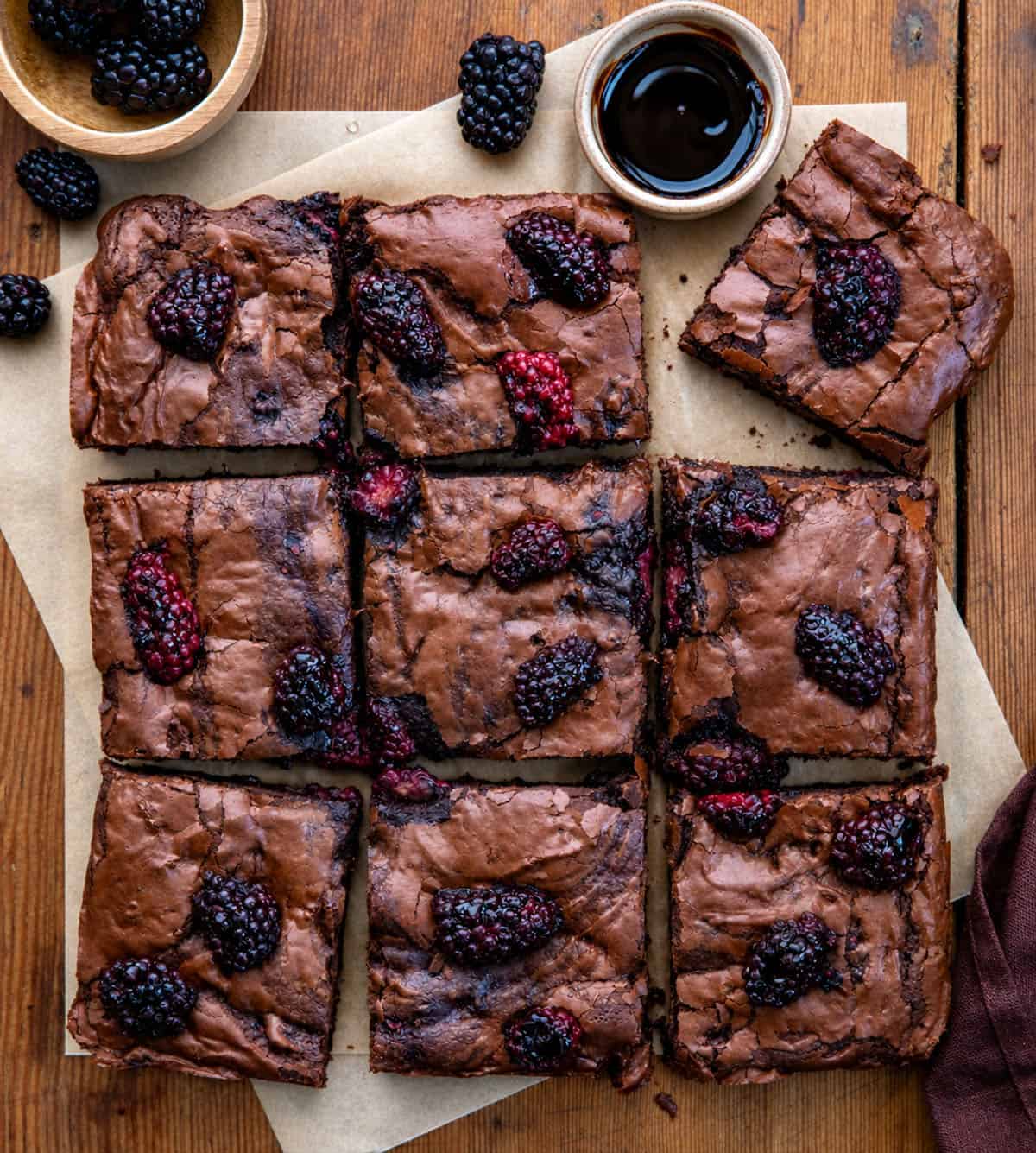 Blackberry Balsamic Brownies cut into squares on a wooden table surrounded by blackberries and balsamic glaze Blackberry Balsamic Brownies cut into squares on a wooden table surrounded by blackberries and balsamic glaze.