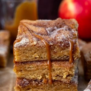 Stack of Caramel Apple Cider Blondies on a wooden table surrounded by caramel, apples, and more blondies.