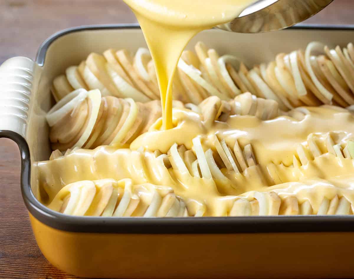 Pouring cheese sauce over raw potatoes and onions in a pan.