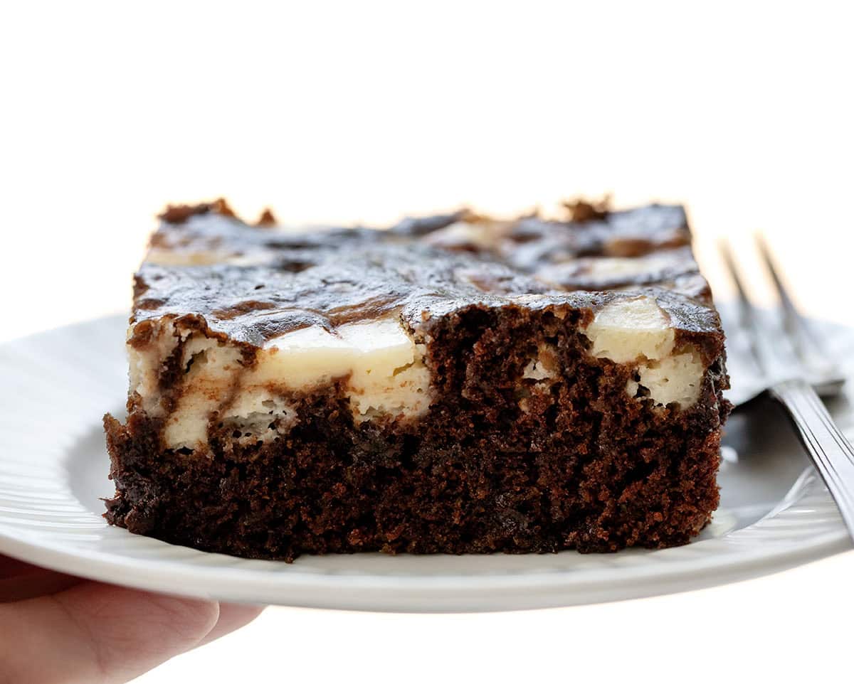 Close up of hand holding a white plate with Cream Cheese Chocolate Zucchini Cake on it.
