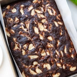Chocolate Zucchini Cake with Cream Cheese Swirl in the pan on a white table with zucchini.