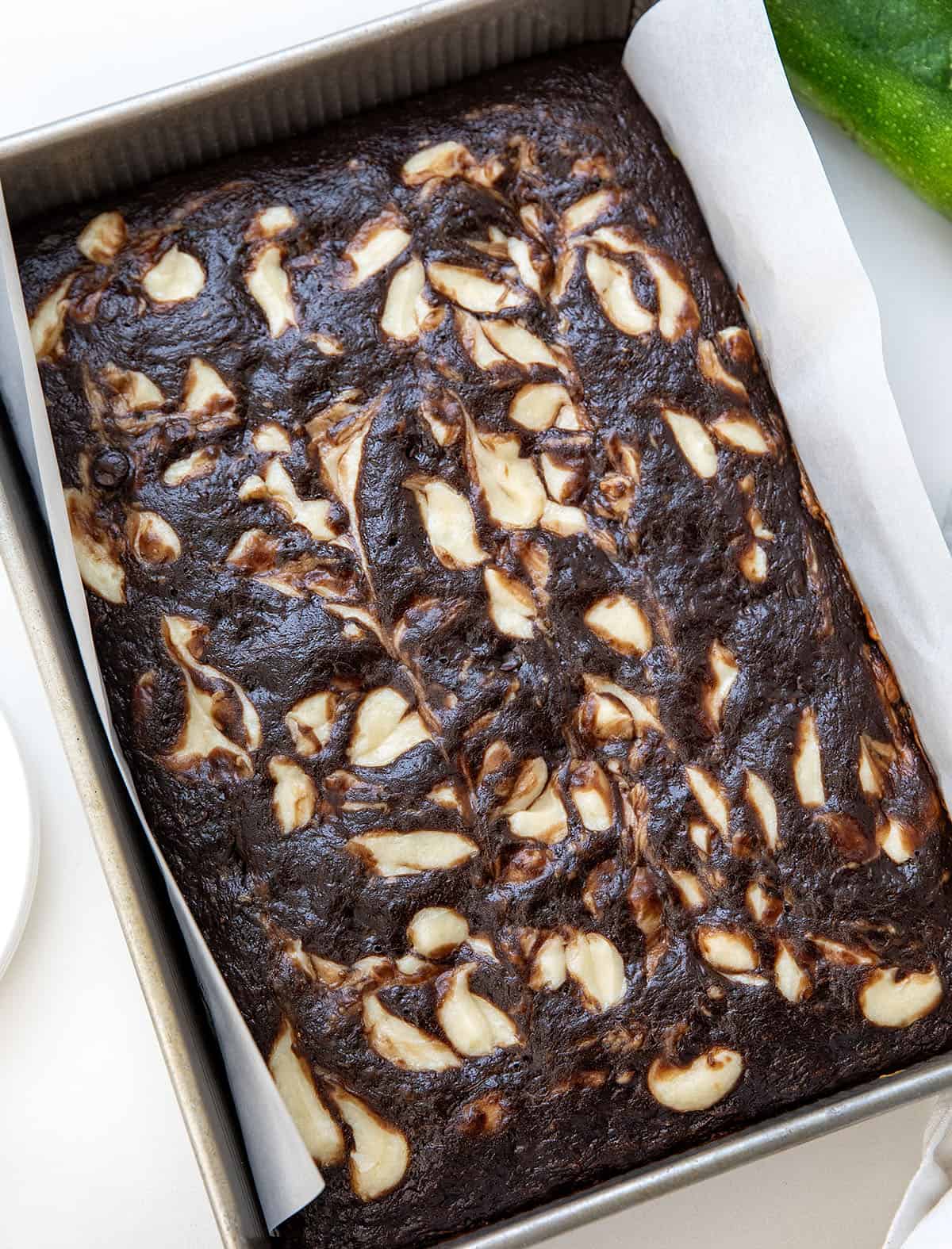 Chocolate Zucchini Cake with Cream Cheese Swirl in the pan on a white table with zucchini.