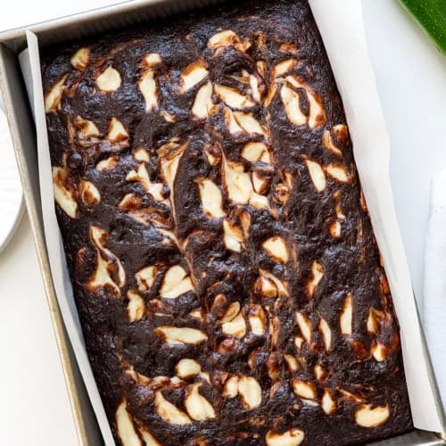 Chocolate Zucchini Cake with Cream Cheese Swirl in the pan on a white table with zucchini.