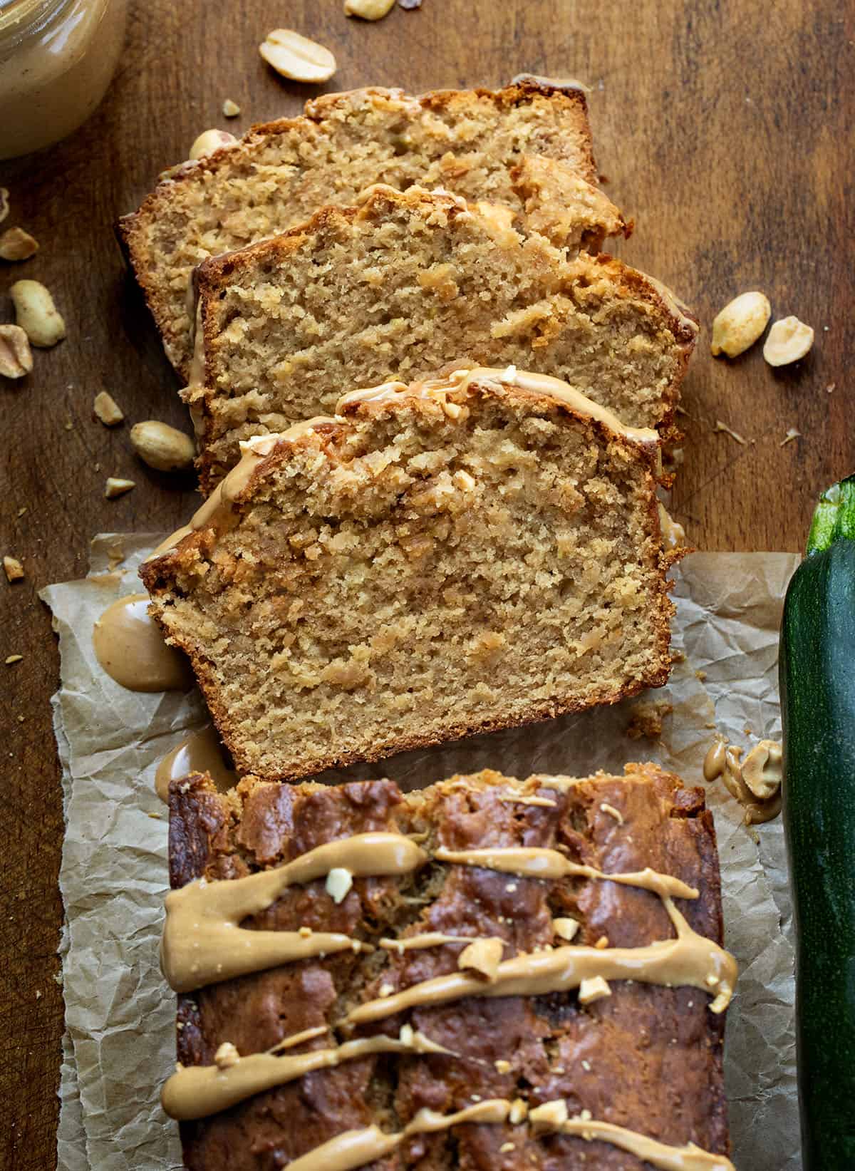 Loaf of Peanut Butter Zucchini Bread on a wooden table with a few pieces cut and laying flat near zucchini and peanuts.