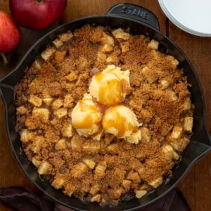 Skillet Apple Brown Betty on a wooden table with white plates and apples from overhead.