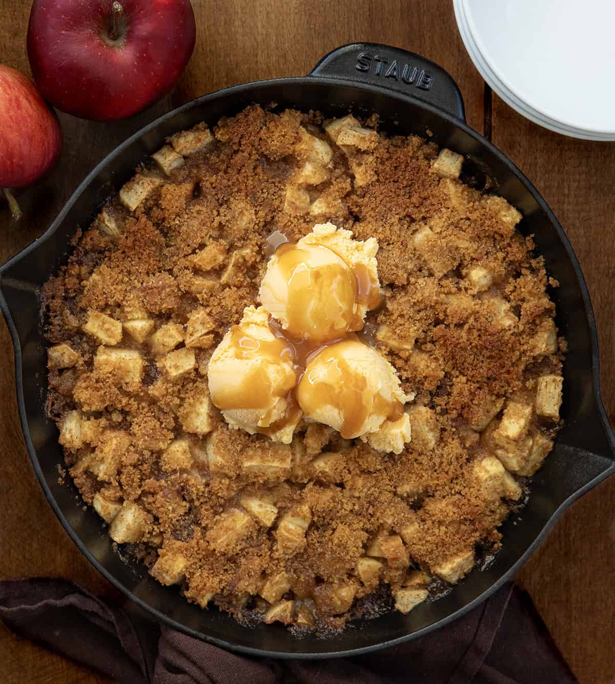 Skillet Apple Brown Betty on a wooden table with white plates and apples from overhead.