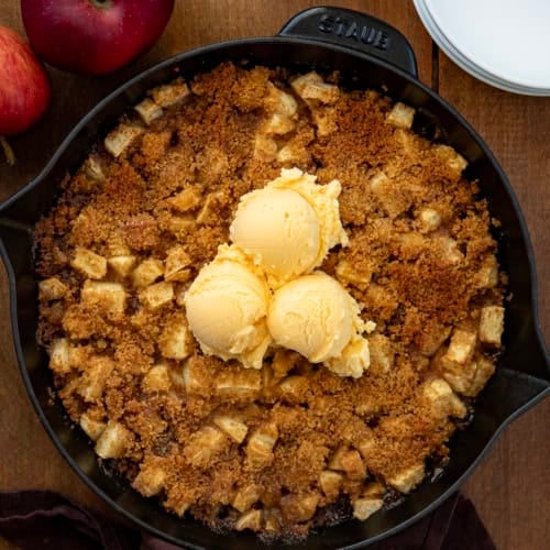Skillet Apple Brown Betty on a wooden table with white plates and apples from overhead.