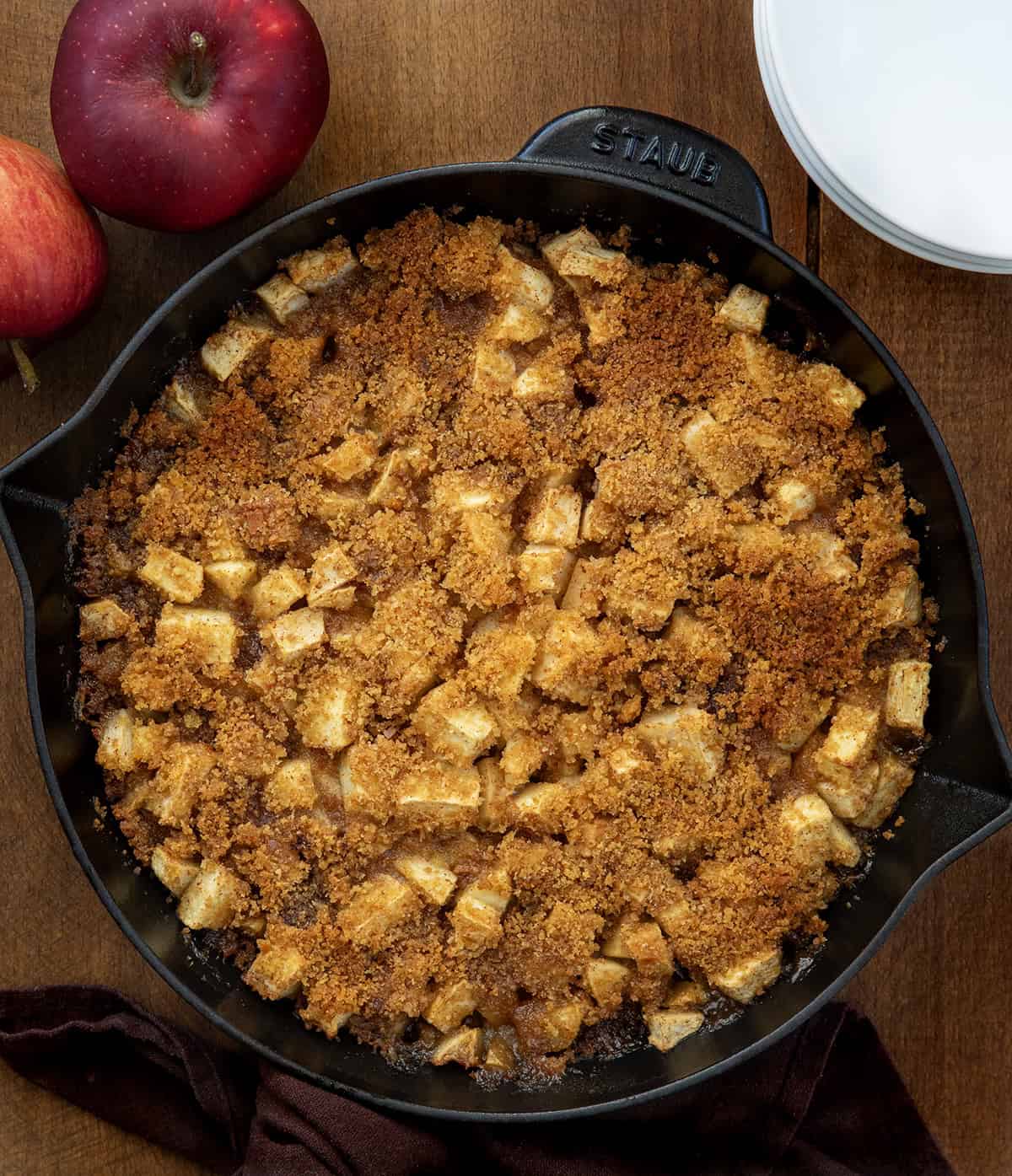 Skillet Apple Brown Betty on a wooden table with white plates and apples from overhead Skillet Apple Brown Betty on a wooden table with white plates and apples from overhead.