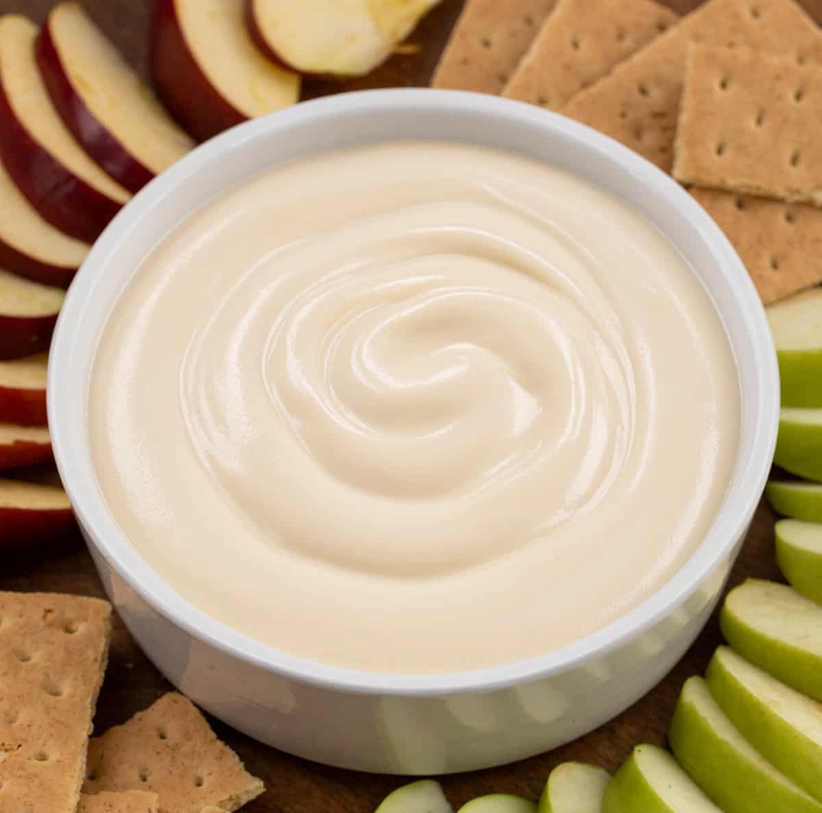 Bowl filled with Apple Cider Marshmallow Sauce surrounded by graham crackers and sliced apples.