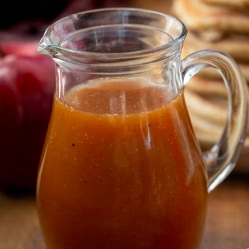 Small glass jar of Apple Cider Syrup on a wooden table with pancakes in the background.
