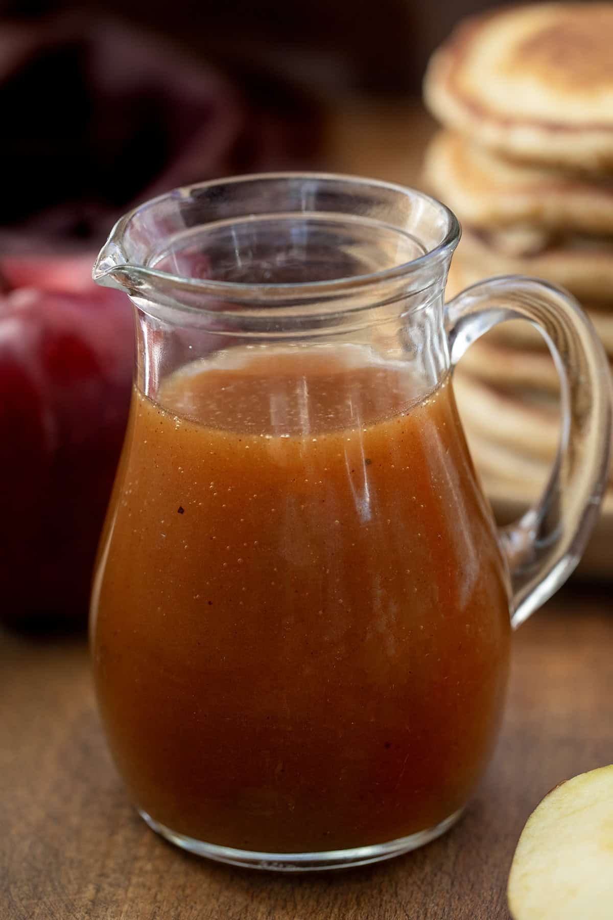 Small glass jar of Apple Cider Syrup on a wooden table with pancakes in the background.