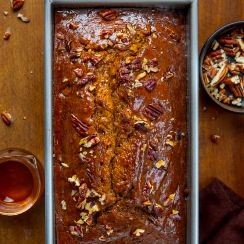 Loaf of Maple Pecan Pumpkin Bread on a wooden table surrounded by syrup and pecans from overhead.