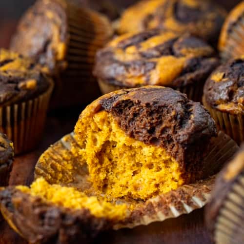 Pumpkin Chocolate Marble Muffins on a wooden table with one muffin halved showing the inside texture.