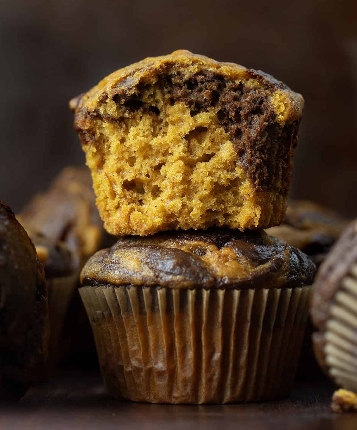 Pumpkin Chocolate Marble Muffins stacked on a wooden table with the top muffin halved showing the inside texture.