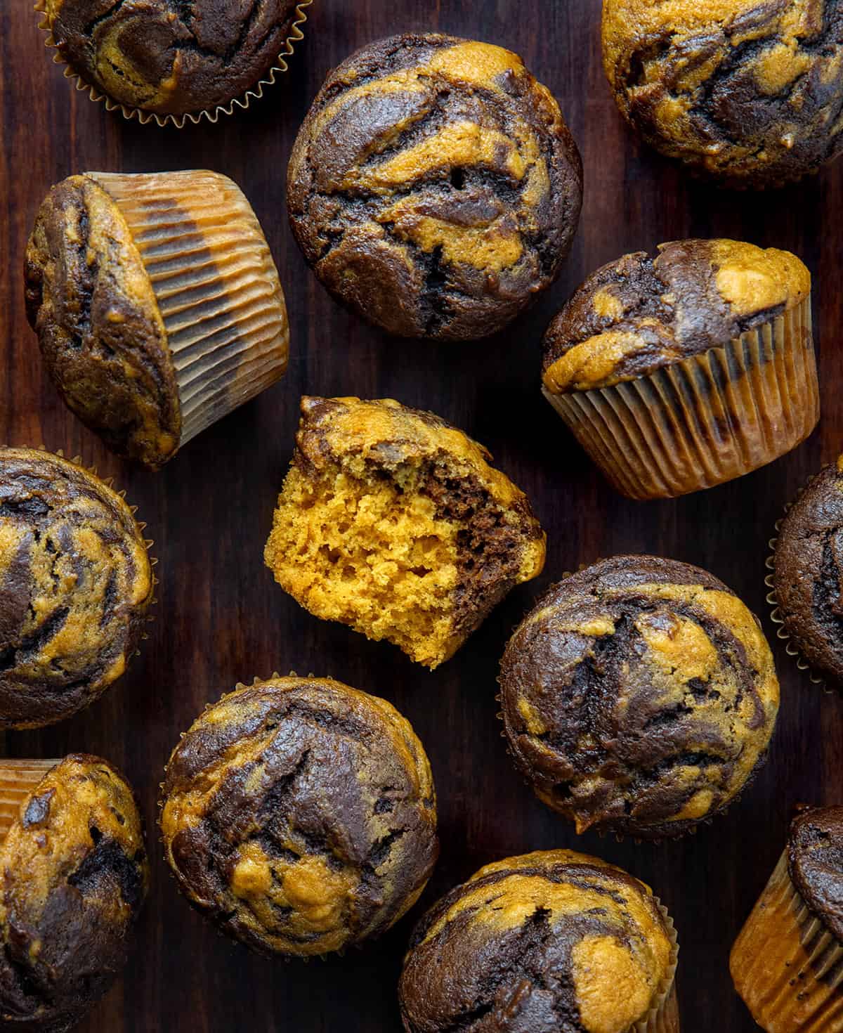 Scattered Pumpkin Chocolate Marble Muffins on a wooden table with one muffin halved showing inside texture.