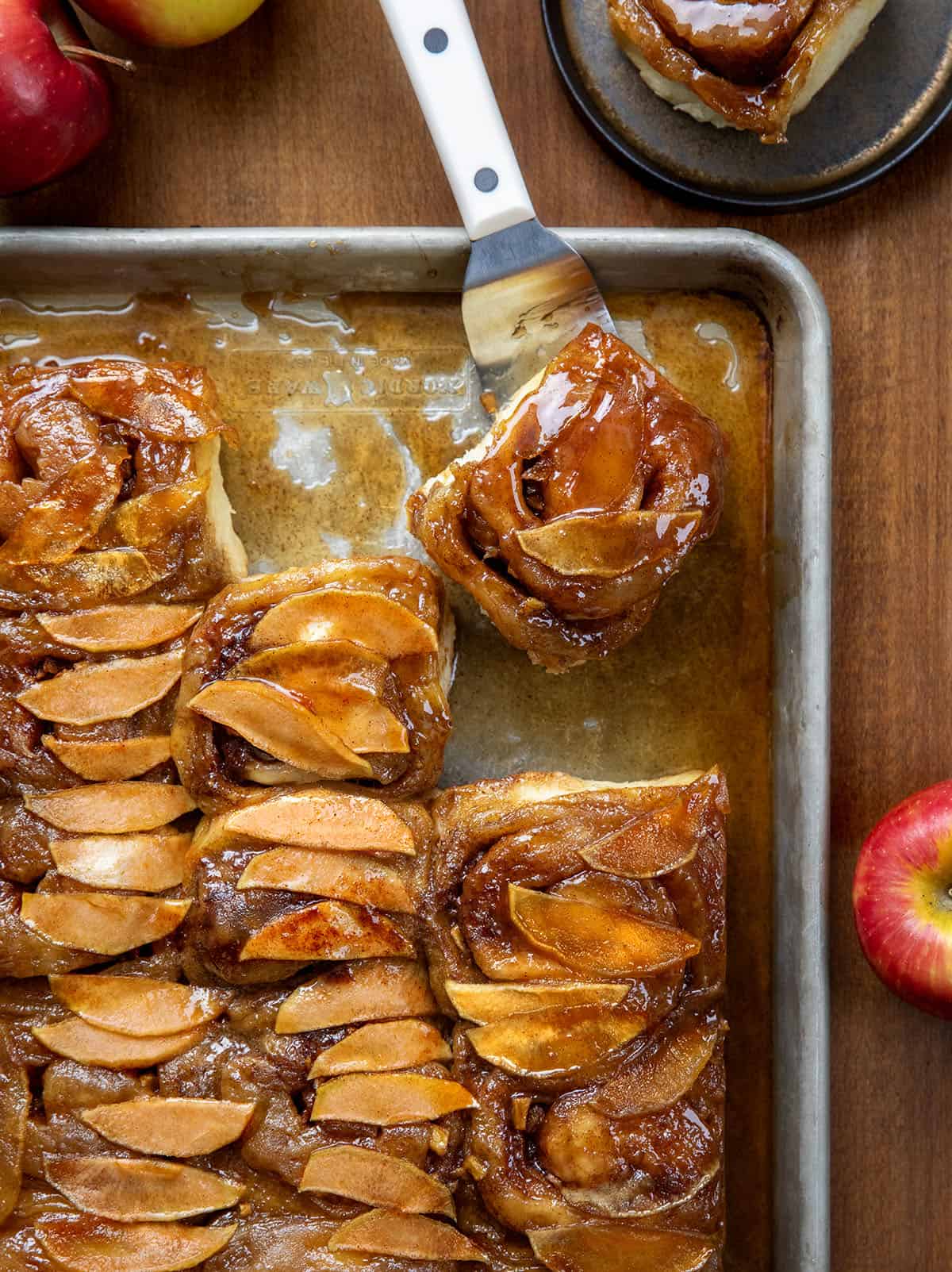 Spatula picking up an Apple Caramel Roll from the pan that is sitting on a wooden table.