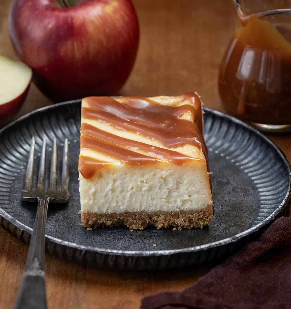 Apple Cider Cheesecake Bar on a plate on a wooden table with apples and apple cider caramel jar.